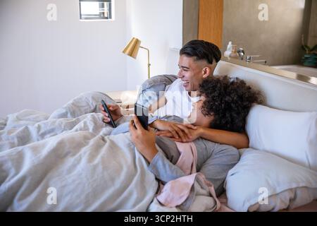 Diverse coppie sdraiate sotto un piumone grigio sul letto, che scorrono sugli smartphone contro la testiera imbottita Foto Stock