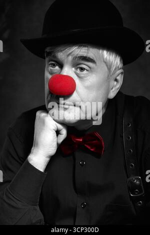 Spettacolare ritratto a colori selettivo di un pagliaccio triste su sfondo scuro. Un uomo anziano con naso rosso da clown e cappello da bowler sulla testa. Vestito con blac Foto Stock