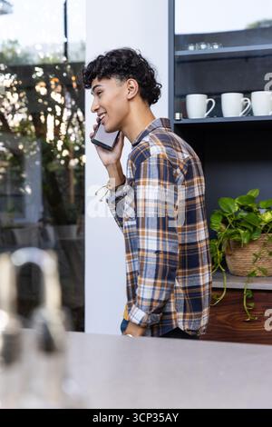 Uomo che parla sullo smartphone accanto al bancone in cucina con tazze bianche, piante, spazio di copia Foto Stock