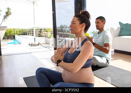 Coppia che pratica yoga sui tappetini a casa tramite porte scorrevoli per la piscina che reggono il peso indossando lo smartwatch Foto Stock