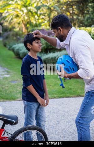 Padre indiano con figlio che regola la cinghia blu del casco da bicicletta accanto alla bicicletta sul sentiero pavimentato del giardino Foto Stock