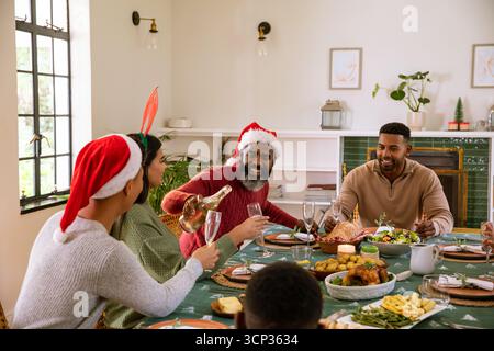 Famiglia variegata nei cappelli di babbo natale al tavolo da pranzo e uomo afroamericano che versa champagne Foto Stock