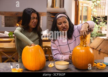 Madre e figlia diverse che raccolgono semi di zucca e polpa dalle zucche sul tavolo da cucina in legno Foto Stock