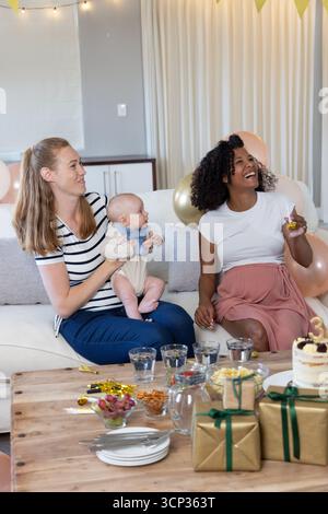 Diverse amiche che festeggiano con il bambino sul divano di casa, torta sul tavolino da caffè Foto Stock