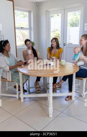 Diverse amiche che sorseggiano dalle tazze intorno al tavolo in sala da pranzo tenendo il bambino in un bavaglino Foto Stock