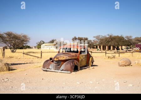 Il cimitero automobilistico di Solitario in Namibia presenta auto d'epoca arrugginite vicino a piccoli edifici desertici, adagiate contro dune e montagne, catturando il fascino del deserto Foto Stock
