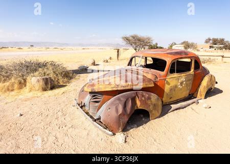 Il cimitero automobilistico di Solitario in Namibia presenta auto d'epoca arrugginite vicino a piccoli edifici desertici, adagiate contro dune e montagne, catturando il fascino del deserto Foto Stock