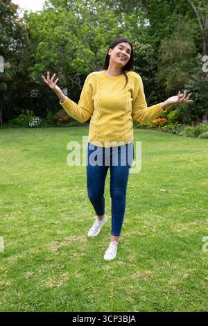 Donna sui trent'anni che stringe le spalle e si allontana sul prato nel cortile posteriore con un maglione Foto Stock
