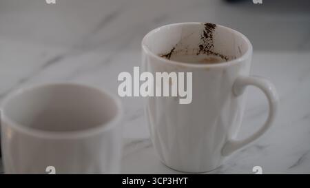 Primo piano di una tazza di caffè in ceramica bianca con macchie di caffè visibili all'interno, posizionata su una superficie in marmo chiaro. Questa immagine è utile per illustrare la conc Foto Stock