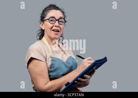 Donna che indossa occhiali e un grembiule che scrive su una clipboard, che gestisce le attività Foto Stock