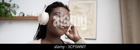 Una donna afroamericana si diverte a trascorrere il tempo in un accogliente appartamento mentre ascolta musica, striscioni Foto Stock