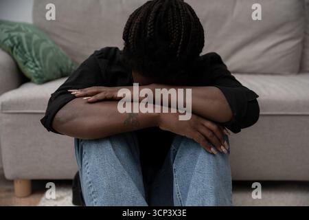 Una donna con i capelli intrecciati si siede sul pavimento, abbracciando le ginocchia in un momento di tristezza a casa. Foto Stock