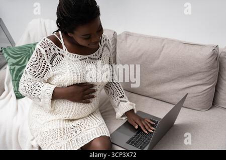 Questa scena accogliente mostra una donna incinta a casa, toccandole dolcemente la pancia mentre usa un notebook. Foto Stock