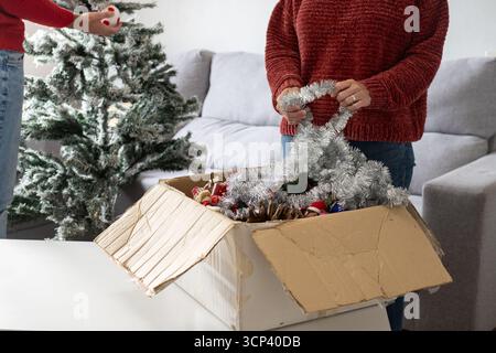 Coppia che disimballa le decorazioni natalizie da una scatola di cartone e inizia a decorare un albero artificiale smerigliato in un accogliente soggiorno. Preparazione per le vacanze Foto Stock