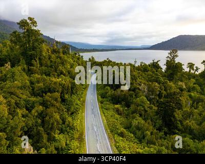 Vista aerea di una strada tortuosa che attraversa una fitta foresta verde che conduce a un lago sereno sotto un cielo morbido e nuvoloso, lago Brunner, costa occidentale, nuova Zelanda. Foto Stock