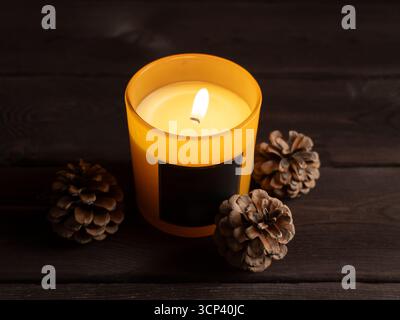 Un colpo dall'alto di una candela profumata di giallo brillante circondata da coni di pino su una superficie di legno scuro. Ambiente caldo e accogliente, perfetto per l'autunno e il Foto Stock