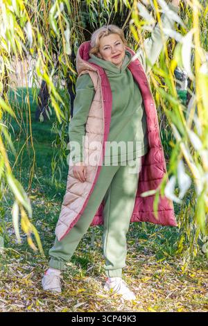 Una donna di mezza età con una tuta verde e un gilet beige e rosa sorridente si erge sulla strada tra i rami appesi di un salice. Brillante Foto Stock