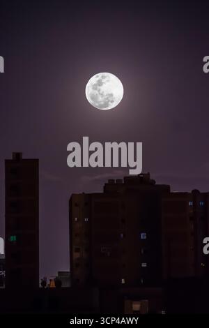 La luna piena splende luminosa nel cielo notturno sopra gli edifici della città, lo skyline urbano sotto la luce della luna in una serata limpida. Foto Stock