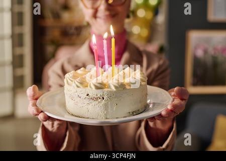 Donna anziana che tiene la torta di compleanno con le candele illuminate che sorridono Foto Stock