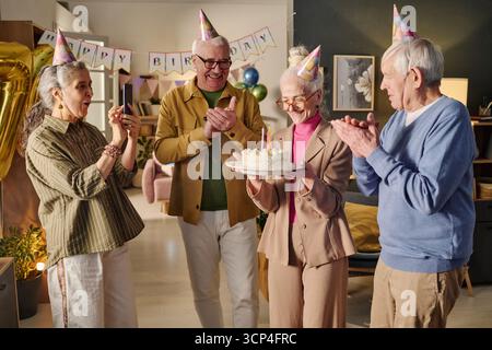 Donna anziana che tiene la torta di compleanno mentre gli amici applaudono sorridendo Foto Stock