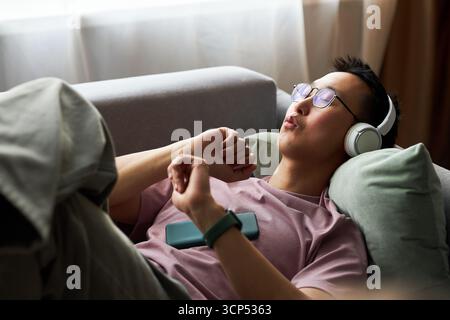 Giovane uomo asiatico che si rilassa sul divano ascoltando musica con le cuffie wireless, gli occhi chiusi, tenendo le mani vicino al viso, lo smartphone si appoggia sul petto e si diverte a divertirsi Foto Stock