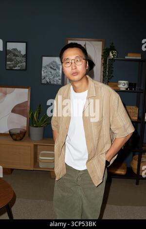 Ritratto di un giovane uomo asiatico adulto in piedi in casa con le mani in tasca, guardando la macchina fotografica con occhiali capelli corti espressione neutra arredamento moderno sullo sfondo Foto Stock