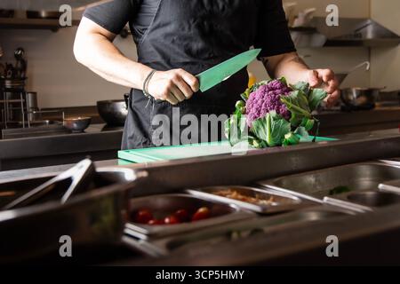 Chef professionista che affetta il vibrante cavolfiore viola su un tagliere in una cucina moderna: Ingredienti freschi, cucina sana e cucina gourmet Foto Stock