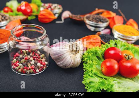 Spezie luminose con curcuma, vari peperoni, sesamo, chiodi di garofano e verdure, pomodori ciliegini, aglio, filis e cucchiai di legno su sfondo nero. Foto Stock