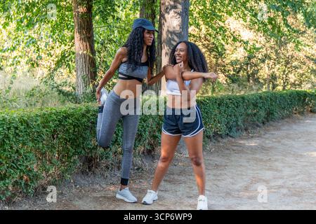 Due donne latine sorridenti che si allungano le gambe e le braccia prima di correre in un parco durante una giornata estiva di sole, promuovendo uno stile di vita sano e il benessere Foto Stock