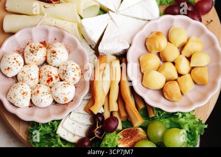 Vassoio di formaggi con prodotti lattiero-caseari assortiti. Palline di mozzarella, formaggio giallo, brie soffice e uva su tavola di legno. Selezione di antipasti gourmet. Foto Stock