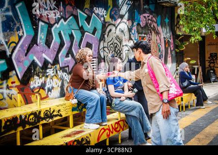 Un gruppo di giovani asiatici si siede e interagisce di fronte a un muro coperto di graffiti su una strada della città, arte e stile di vita urbano di kuala lumpur, malesia Foto Stock