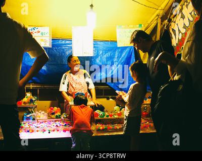 TOKYO, GIAPPONE - 20 settembre 2025: Un venditore sorridente interagisce con i bambini che giocano a un gioco in uno stallo notturno matsuri estivo Foto Stock