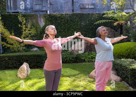 Diverse amiche in abbigliamento sportivo che praticano yoga stretch su prato con tappetini e pareti ricoperte di edera Foto Stock