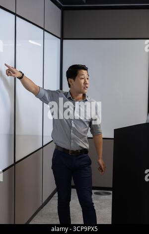 Uomo asiatico che indossa una camicia button-up presentando al leggio nero con pannelli bianchi nella sala conferenze Foto Stock