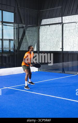 Uomo afro-americano con racchetta in padel, palla su campo blu con pareti in vetro e linee bianche Foto Stock