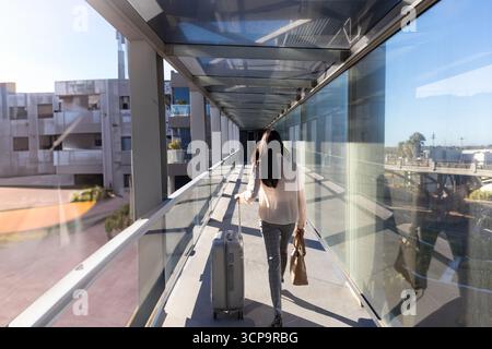 Donna asiatica che cammina attraverso il ponte sospeso tirando la valigia rotolante e trasportando una borsa tote in pelle Foto Stock