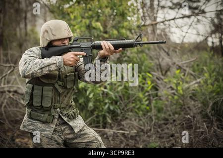 Soldato maschio inginocchiato, fucile mirato e indossa giubbotto e casco mimetico nella foresta, copia spazio Foto Stock