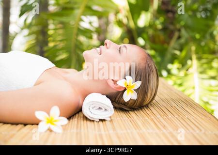 Donna sdraiata su un tavolo da massaggio in bambù all'aperto presso il centro benessere avvolto in un asciugamano con fiore di frangipani Foto Stock