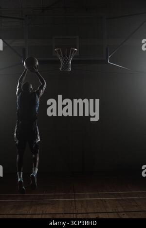 Pallacanestro che vola verso il cerchio in palestra poco illuminata, con luci spot che proiettano ombre spettacolari Foto Stock