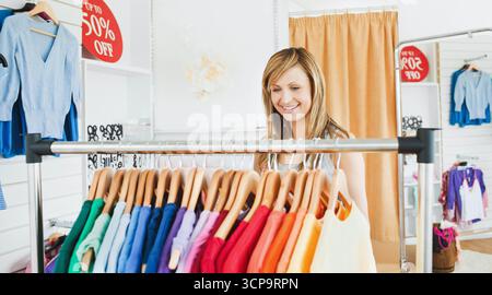 Donna shopper che sfoggia camicie su grucce di legno in boutique store con cartello rosso di vendita e spazio copia Foto Stock