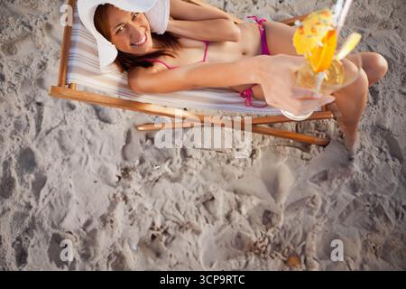 Donna sdraiata su una sedia da spiaggia con bikini rosa e cappello che si estende su un bicchiere da cocktail con ombrellone Foto Stock