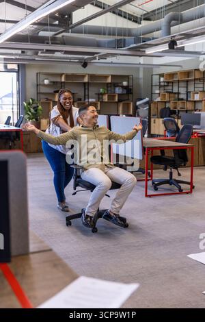 Diversi colleghi spingono la sedia da ufficio su ruote lungo il corridoio in ufficio mentre ridevano Foto Stock