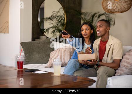 Coppia afroamericana seduta sul divano nel soggiorno, guardando la TV con telecomando e mangiando da asporto Foto Stock