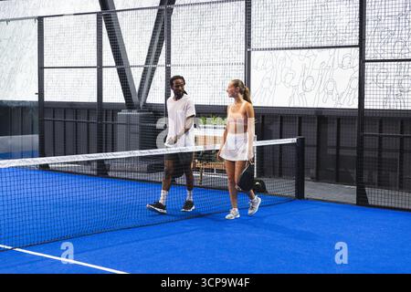 Diversi partner sportivi che camminano sul campo in padel blu sotto la rete indossando abbigliamento sportivo con racchette Foto Stock