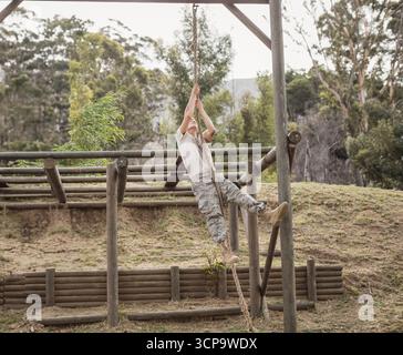 Allievo militare maschile che indossa una corda per arrampicata camo su una pista a ostacoli con telaio di legno in radura erbosa Foto Stock