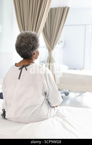 Donna afroamericana anziana seduta sul letto nella stanza d'ospedale che indossa un abito da ospedale accanto alla pompa IV Foto Stock