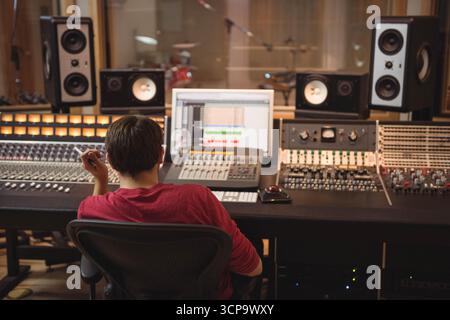 Ingegnere audio maschio che mixa tracce su console nella sala di controllo dello studio con kit batteria in background Foto Stock