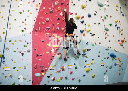 Una donna bianca che scala colorata si tiene sulla parete della palestra di arrampicata utilizzando una borsa di gesso con imbracatura di corda Foto Stock