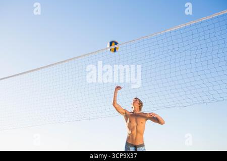 Uomo atletico che indossa pantaloncini da nuoto che giocano a pallavolo su rete sulla spiaggia sotto il cielo blu, copia spazio Foto Stock
