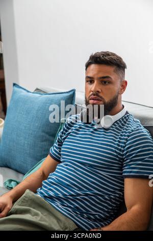 Uomo ispanico seduto sul divano con cuscini blu e cuffie sovrauricolari Foto Stock